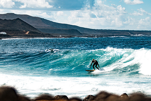 Surfer rijdt op een heldere blauwe golf aan de ruige kust van de Canarische Eilanden, met rotsen, heuvels en een wolkenlucht op de achtergrond.